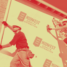 Two workers install building wrap panels featuring the Midwest Supply & Distribution logo on an unfinished building wall.