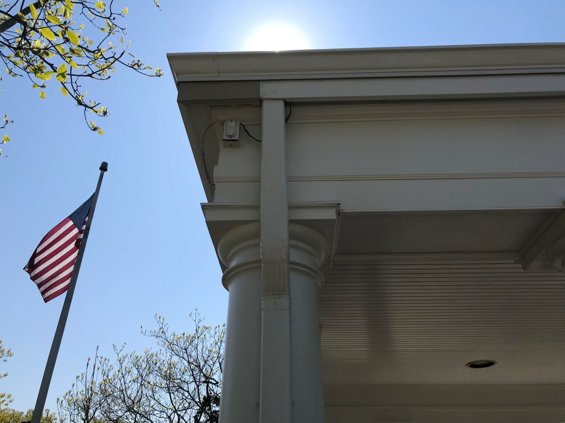 American flag waving next to a white building with classical columns, under a bright sun.
