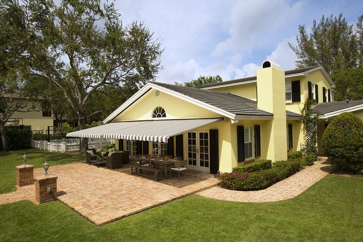 Yellow house with a striped awning over a patio with a dining table, surrounded by a lawn.
