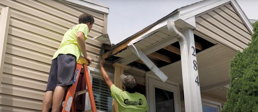 Century Home Improvements - Two people repairing a house's eaves, one on a ladder. House has tan siding and white trim.