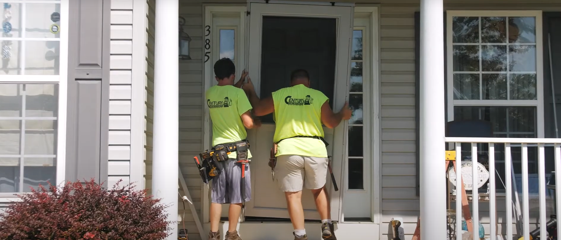 Century Home Improvements - Two workers installing a screen door on a porch of a house.