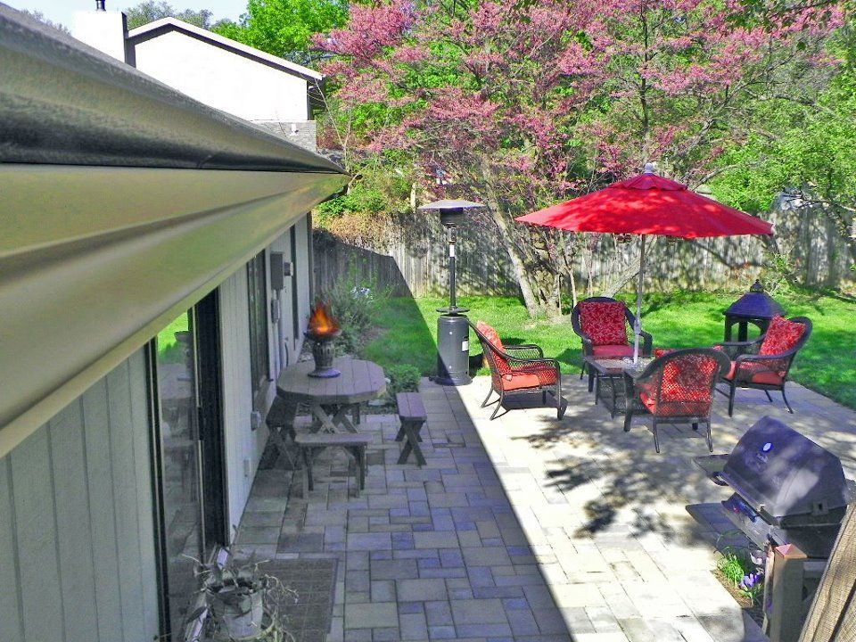 Patio with red umbrella, seating area, grill, brick pavers, and lush green yard.