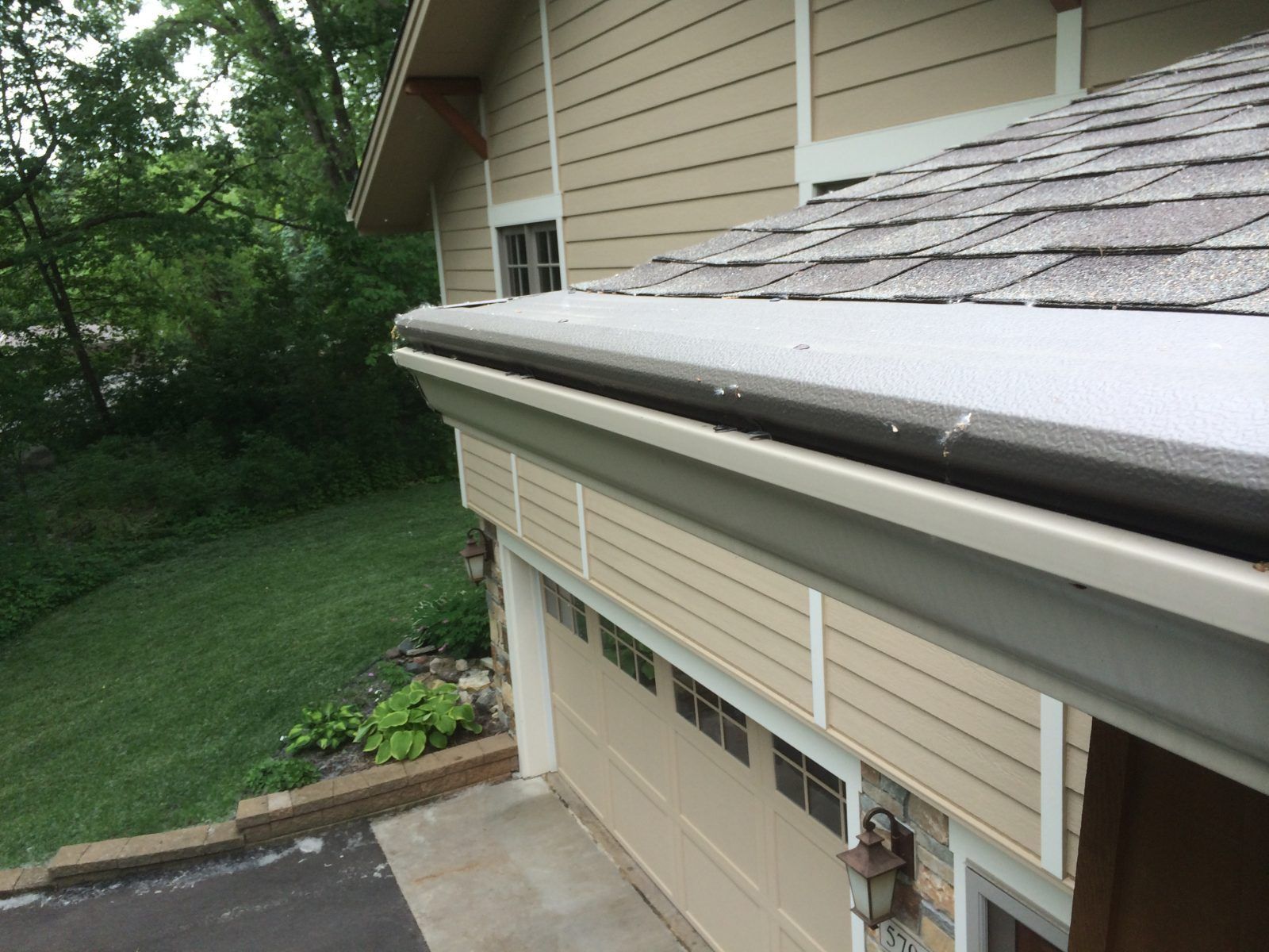 Gutter on a house with tan siding and a dark roof. The gutter is dark, and the garage door is beige.