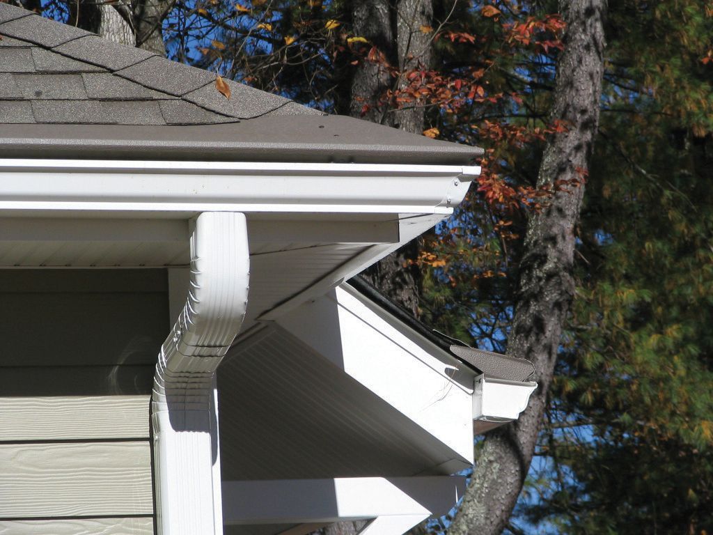 White gutters and eaves on a house with brown shingles and trees in the background.