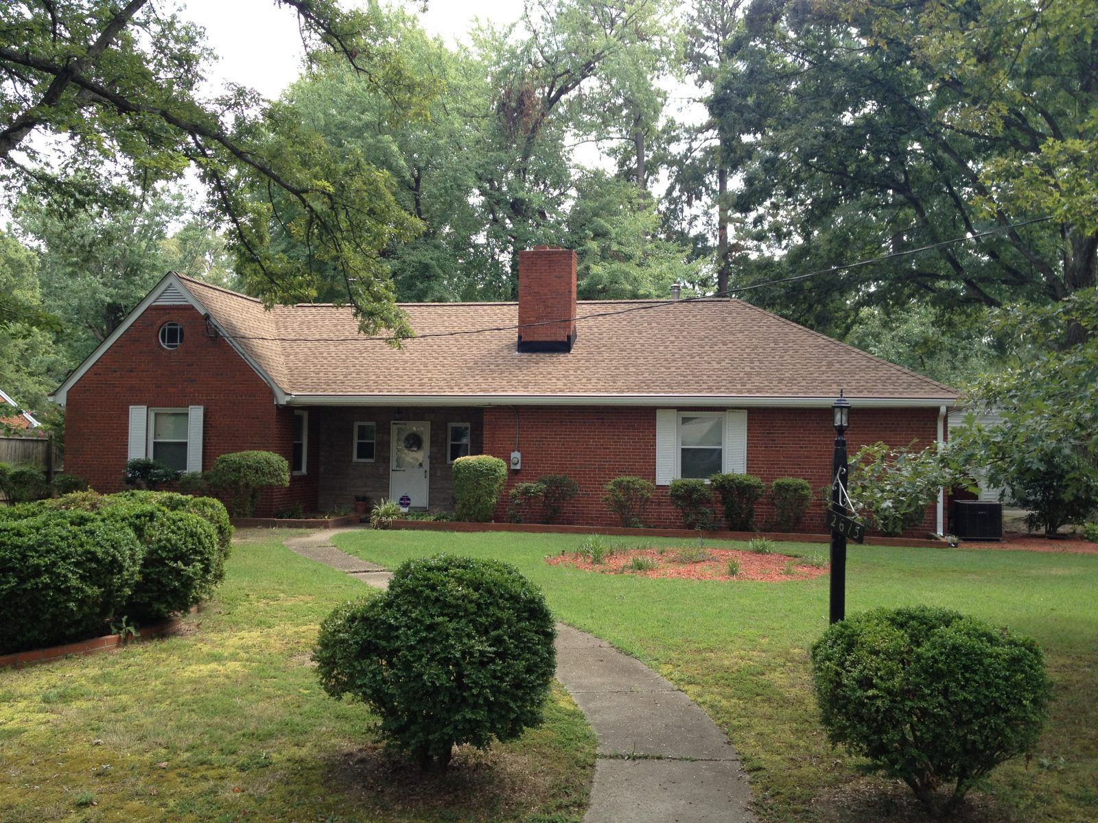 Red brick house with a brown roof, chimney, white windows, and a curved walkway. Lush green bushes and trees surround it.
