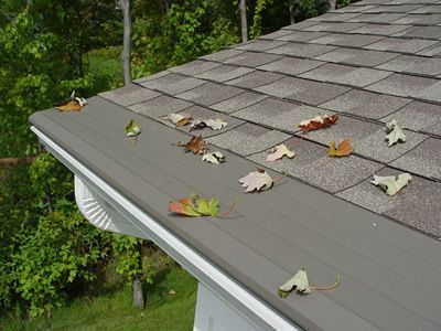 Close-up of a roof with shingles and a gutter filled with fallen leaves.