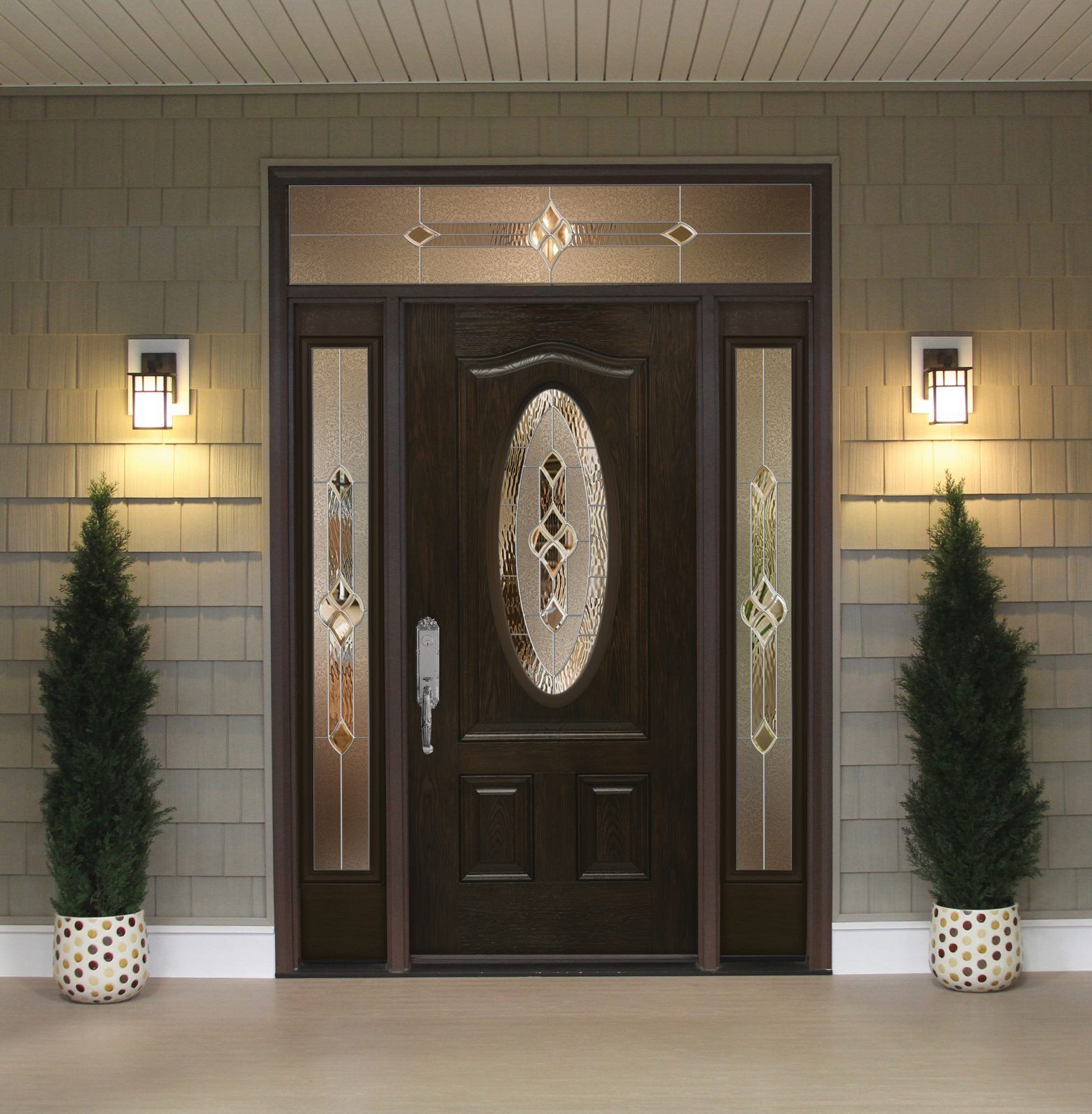 Dark brown front door with oval glass, sidelights, and transom, flanked by potted evergreens.