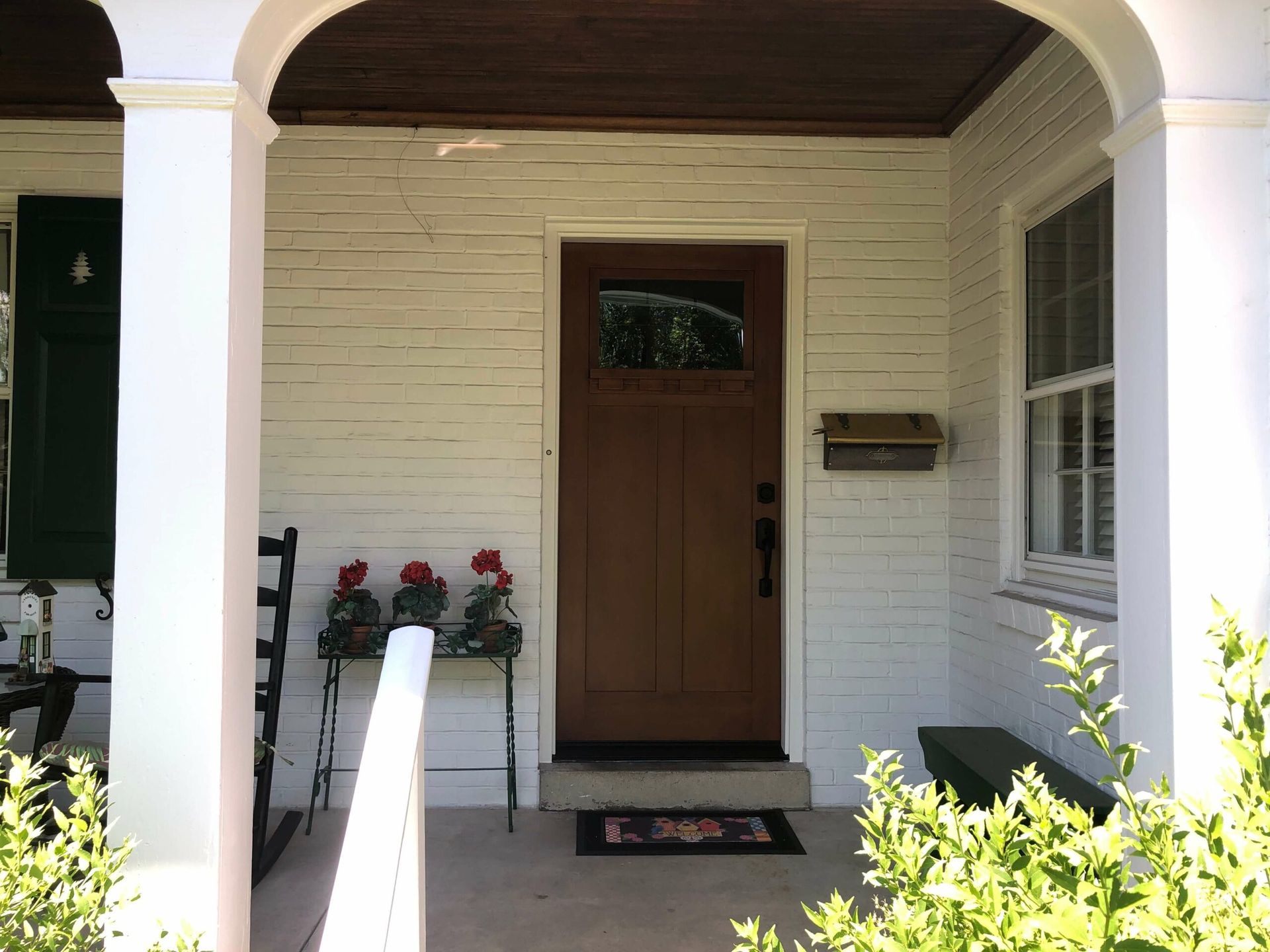 Front porch of a house with white brick walls, a brown door, and a wrought iron plant stand with red flowers.