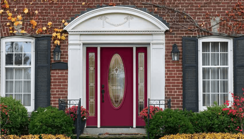 A red door with oval glass and sidelights framed by white trim, flanked by windows with black shutters.