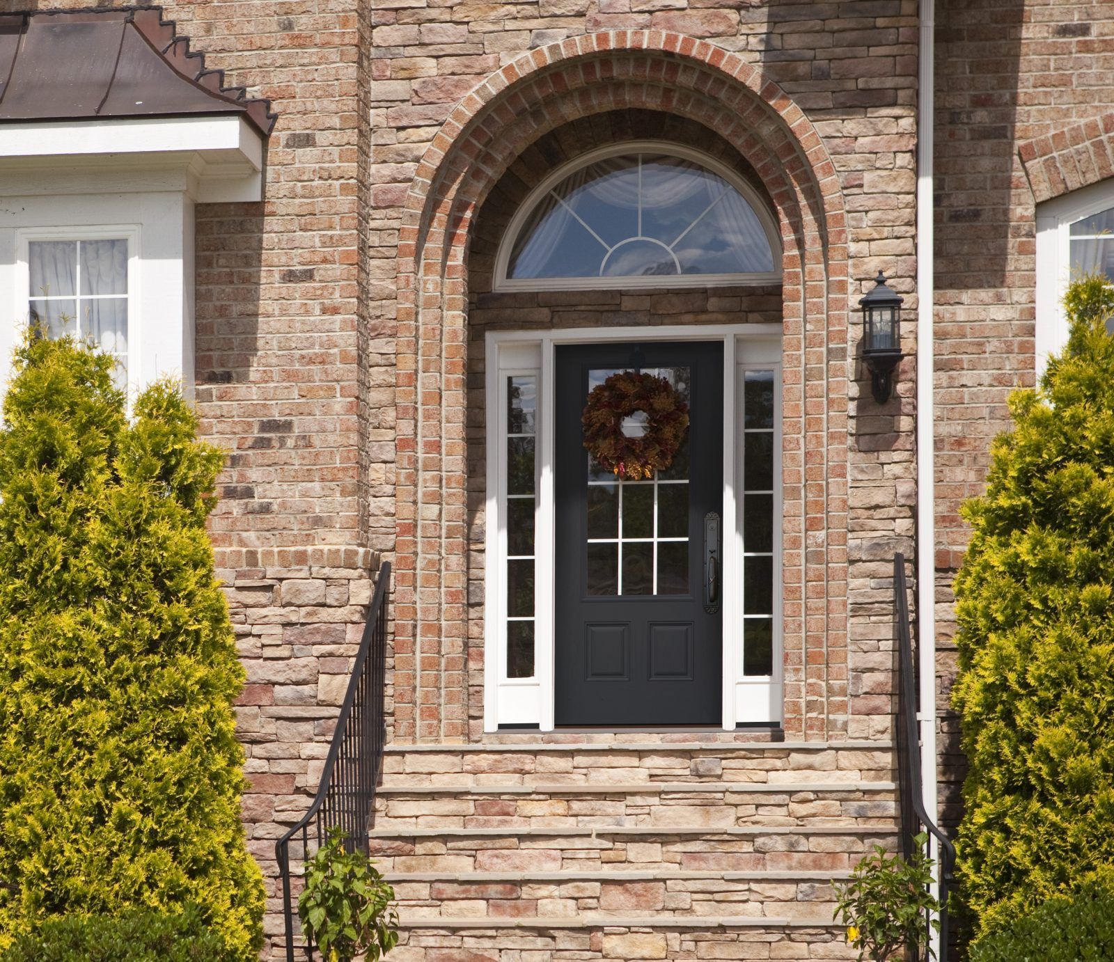 Brick home entrance with black door, arched window, and steps. A fall wreath hangs on the door.