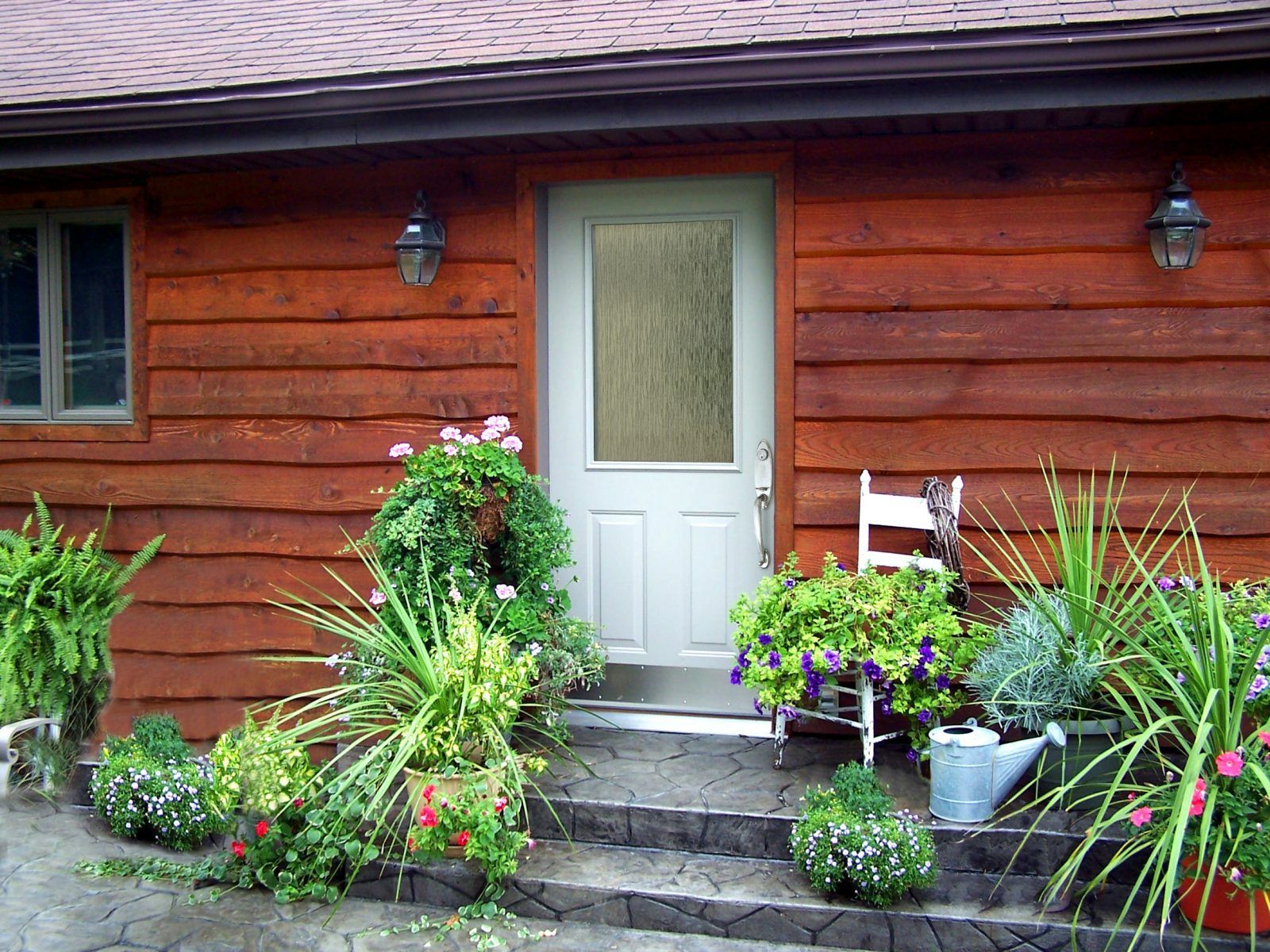 Red wooden cabin with a white door, gray steps, and colorful potted plants. Two outdoor lights flank the door.
