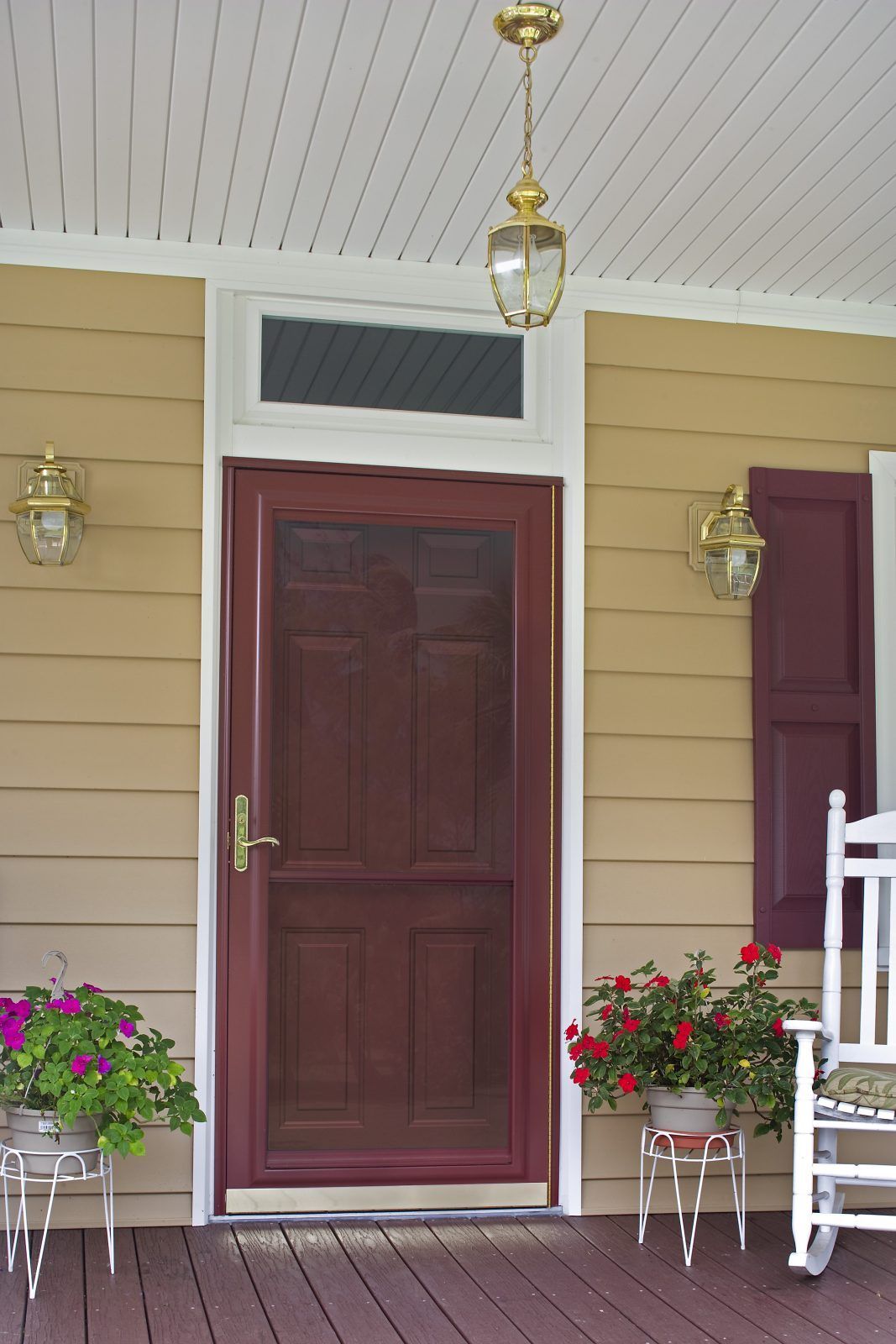 A burgundy front door with screen on a porch, flanked by sconces and potted flowers. 