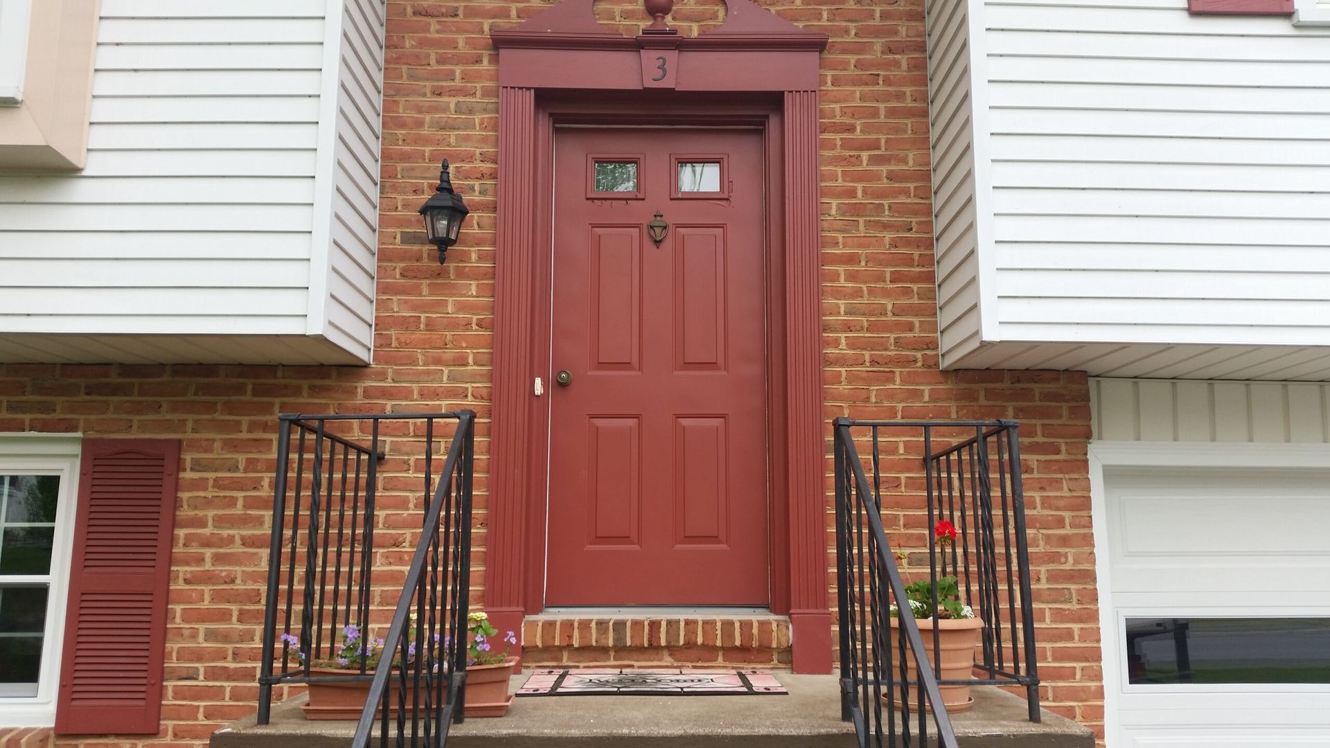 Red front door with brick surround and black iron railings. White siding and a garage door flank the entrance.