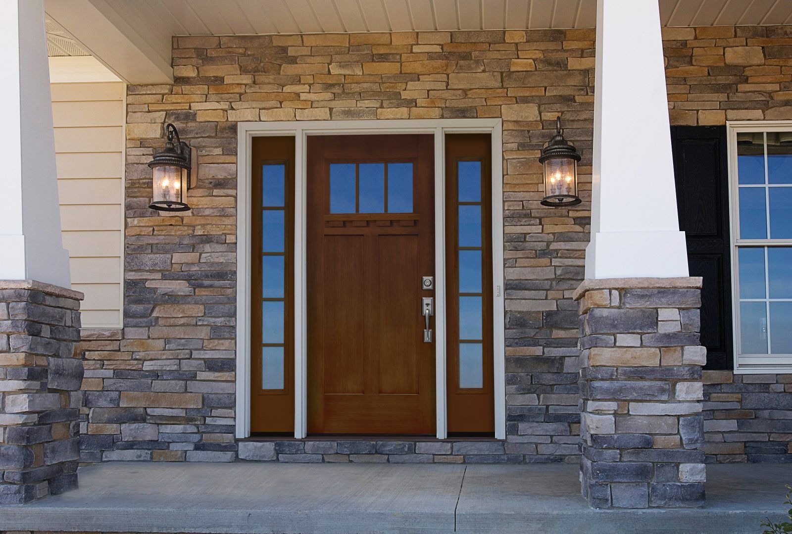 A wooden front door with sidelights and transom window, framed by stone veneer and flanked by porch columns and lanterns.