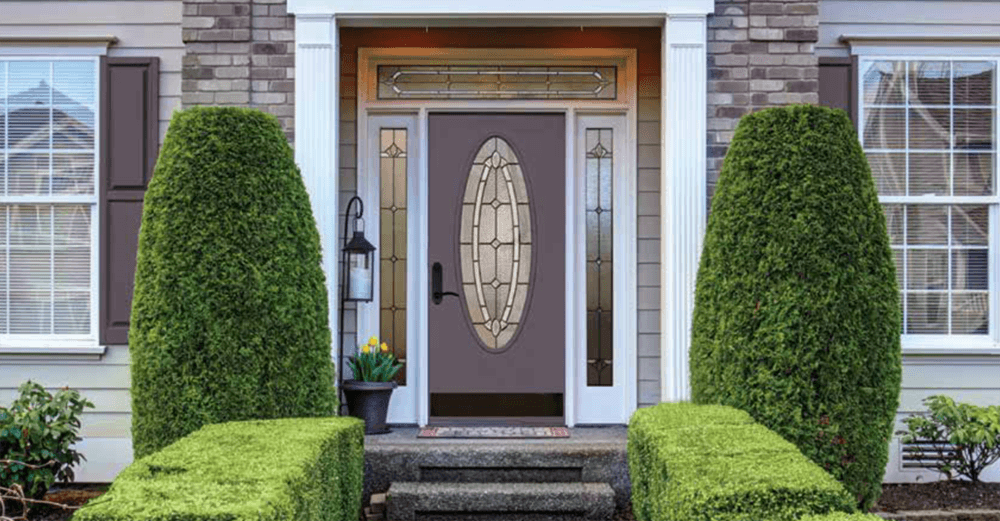 A house's front entrance with a gray door, oval window, and two symmetrical green bushes. 