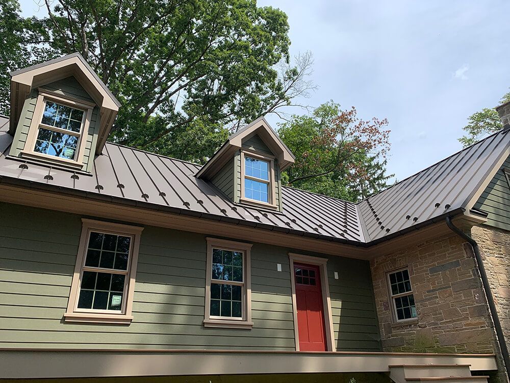 Green house with metal roof, dormers, and red door, surrounded by trees.