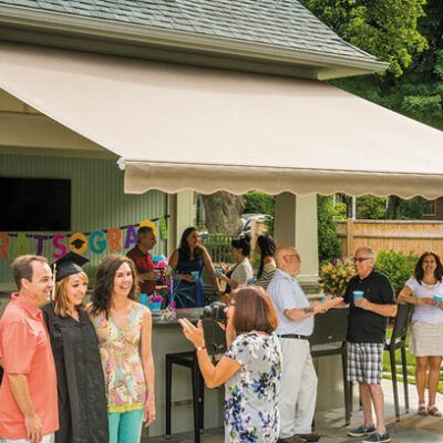 Graduation party outdoors: people gathered, posing for photo in front of a bar, awning overhead.