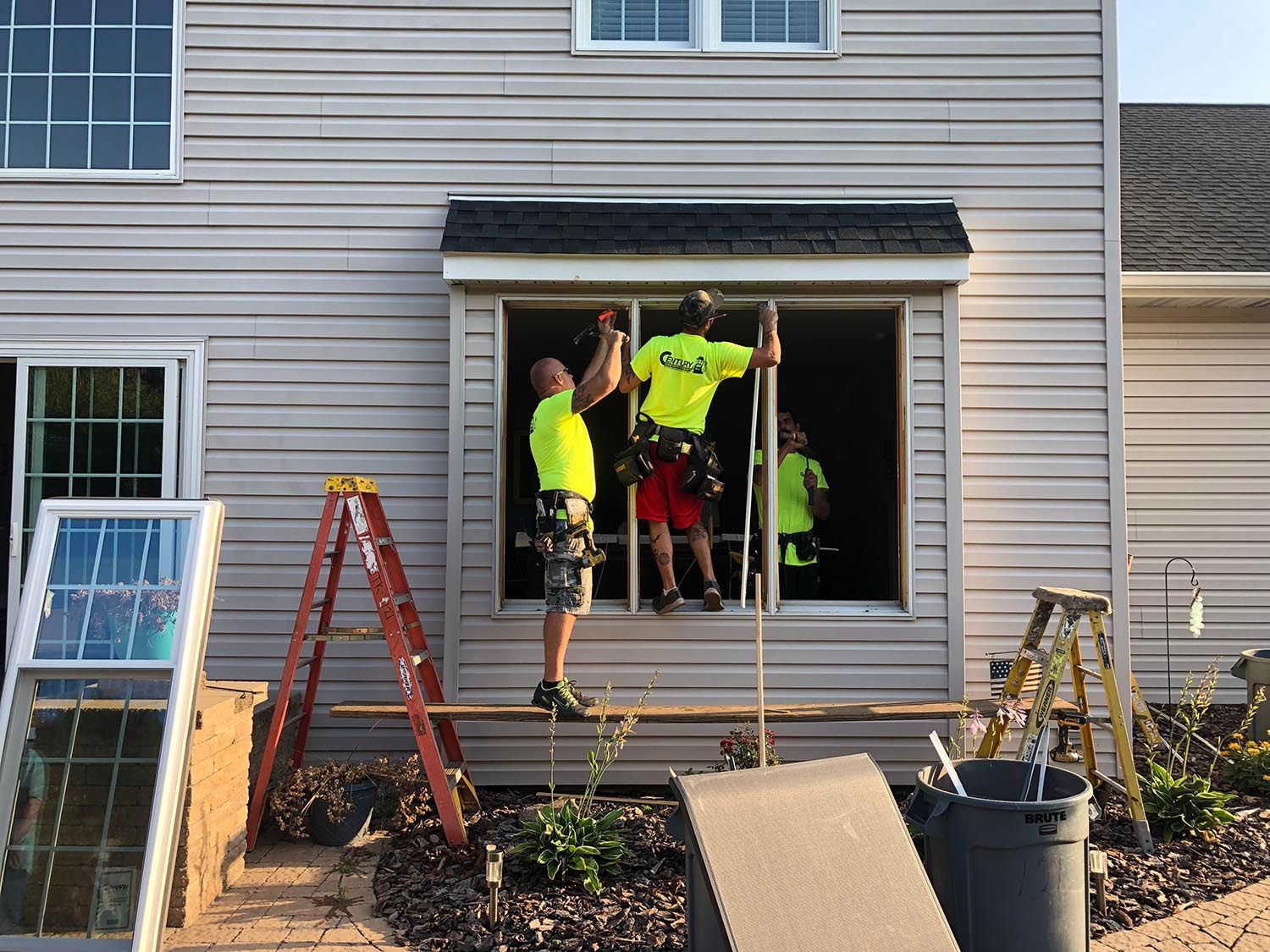 Three workers installing a window, standing in the opening. Beige house with installed and uninstalled windows nearby.