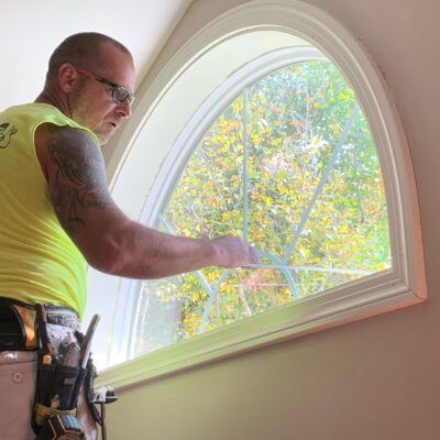 Man cleaning a large arched window with a scraper, wearing a yellow shirt and tool belt, indoors.