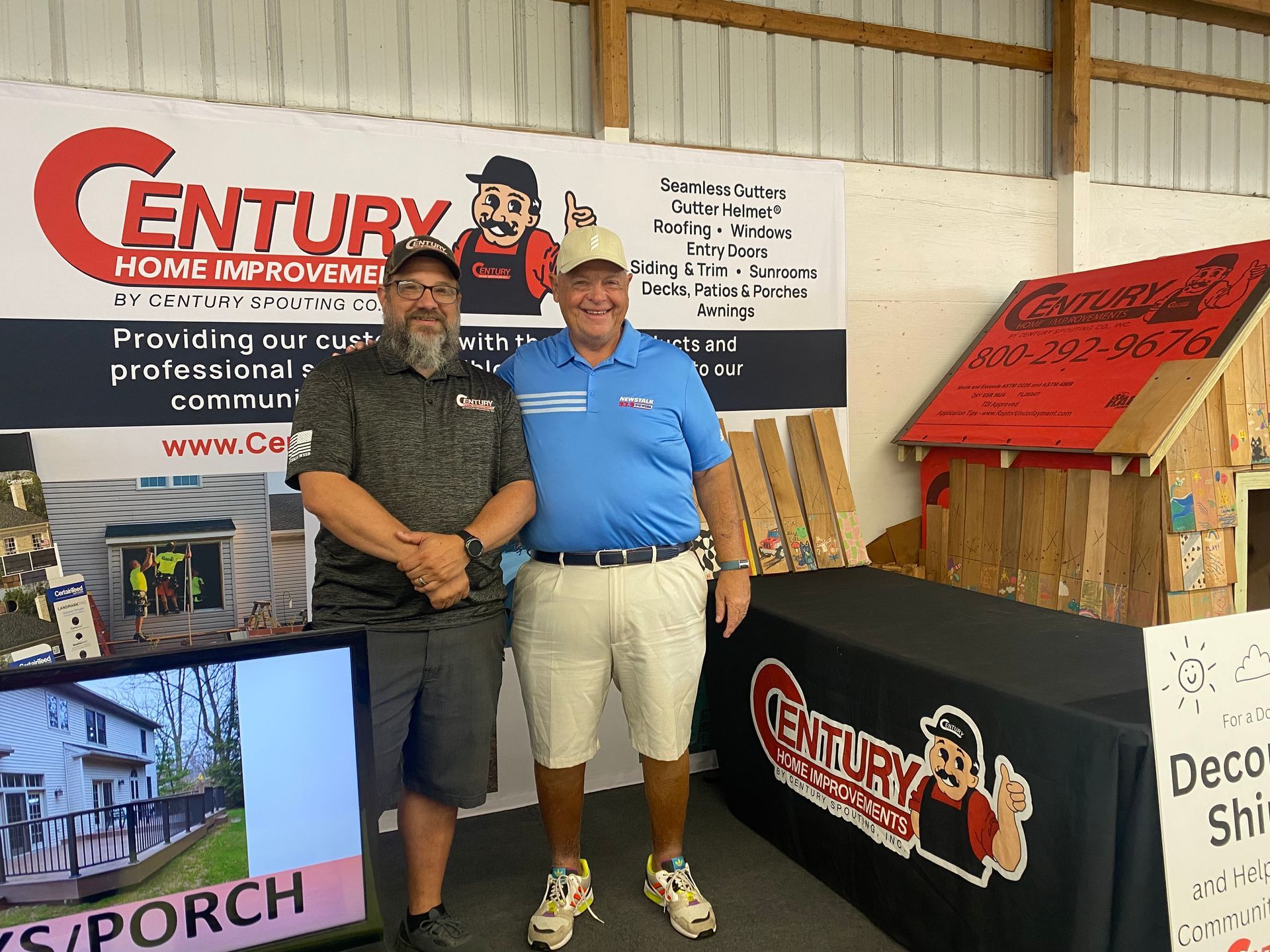 Two men stand in front of a Century Home Improvements booth, smiling. There's a small house model and a banner.