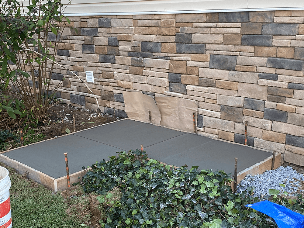 Freshly poured concrete patio with wooden borders, set against a stone wall, in a yard.