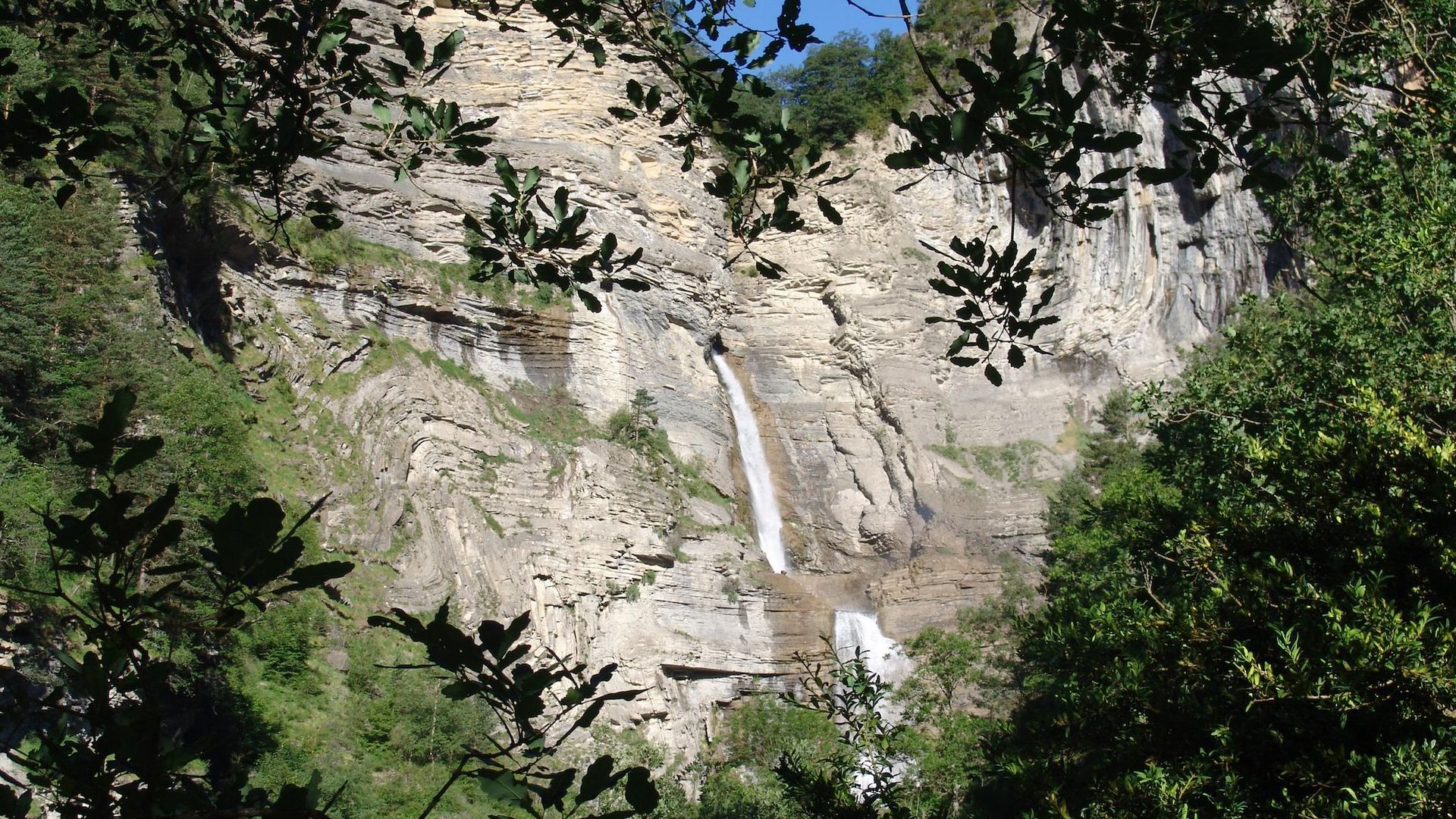Cascada que cae por un acantilado rocoso, enmarcada por follaje verde bajo un cielo soleado.