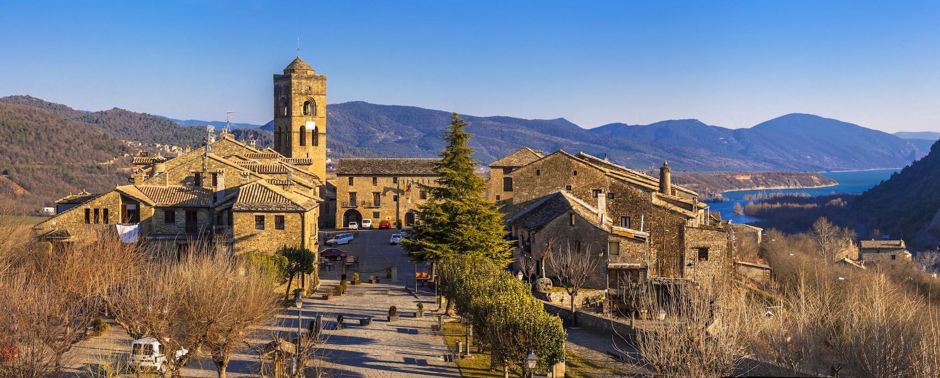 Un pueblo medieval de piedra en la cima de una colina, con una torre alta. Cielo azul y montañas al fondo.