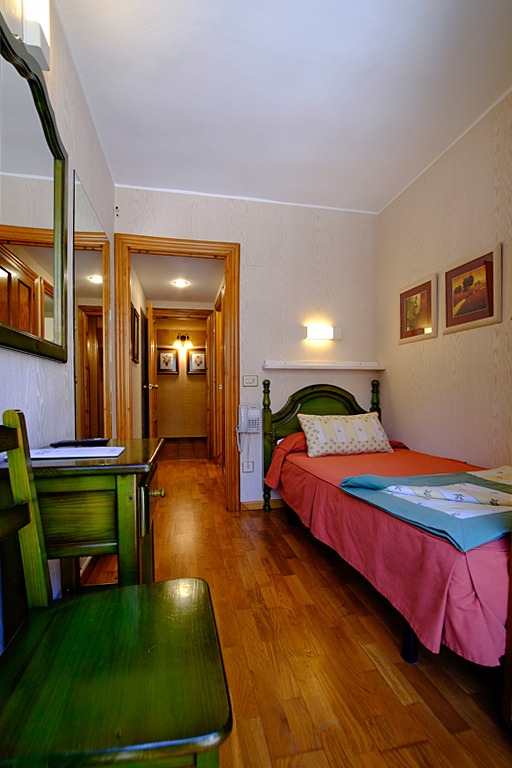 Pequeña habitación de hotel con una cama individual, escritorio y un pasillo que conduce a otras habitaciones.