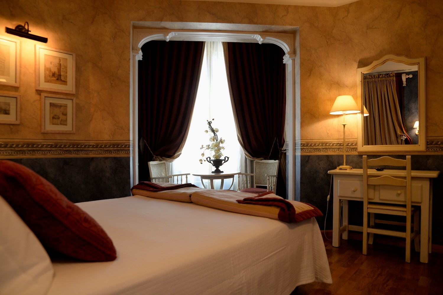 Acogedora habitación de hotel con una cama blanca, detalles en color burdeos y una ventana con cortinas oscuras.