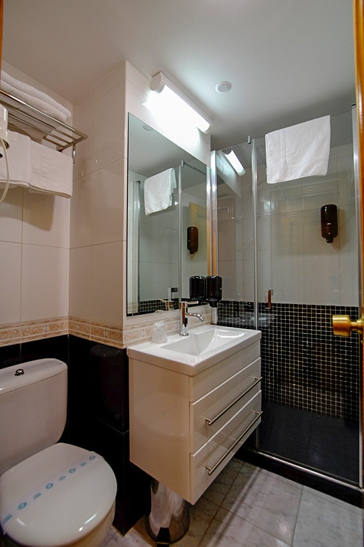 Baño pequeño con inodoro, lavabo y ducha. Interior en blanco, negro y cristal, con toallas.