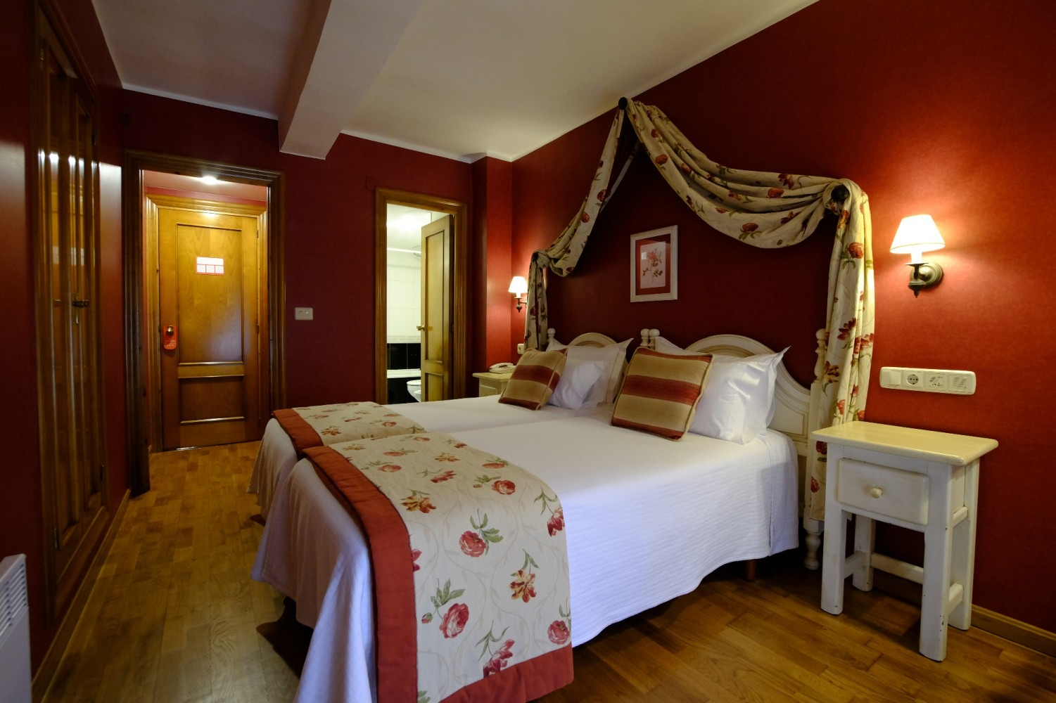 Dormitorio de hotel con paredes rojas, cama doble, camino de cama con motivos florales, suelo de madera, mesita de noche .