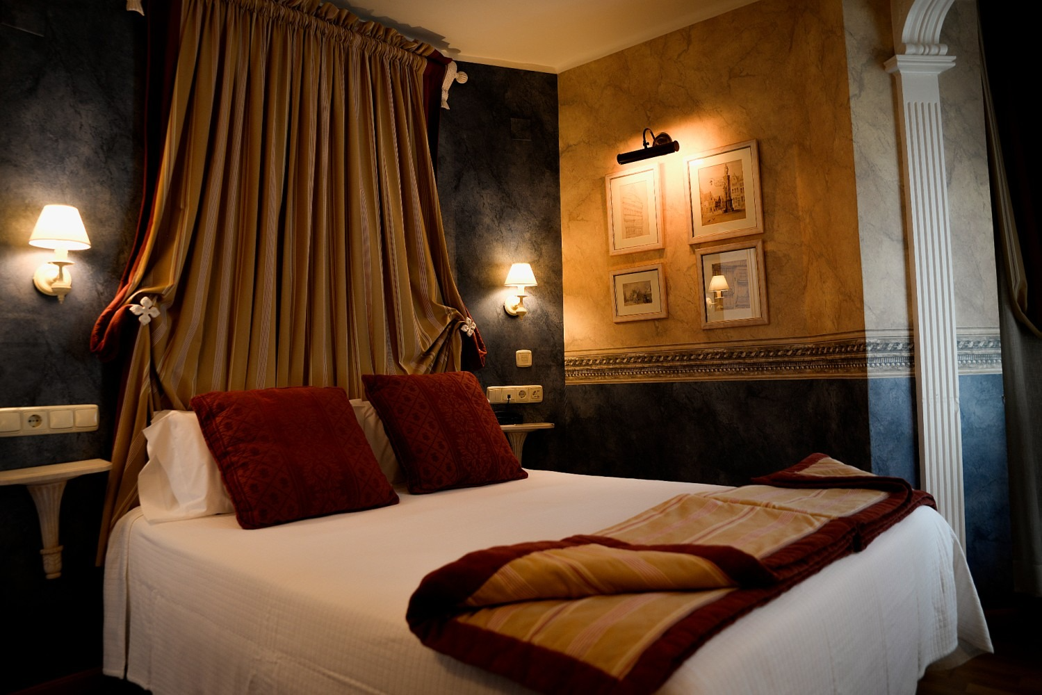 Acogedor dormitorio con una cama, almohadas y manta color burdeos, cortinas doradas, apliques de pared y obras de arte.