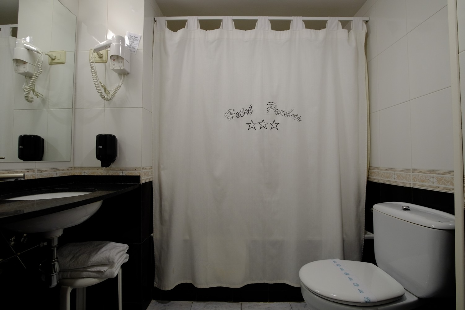 Baño con inodoro, lavabo, espejo, cortina de ducha y secador de pelo en la pared.