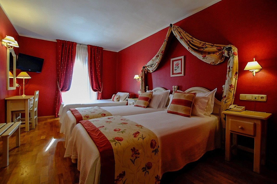 Habitación de hotel con dos camas individuales, paredes rojas, dosel, mesa pequeña y ventana.