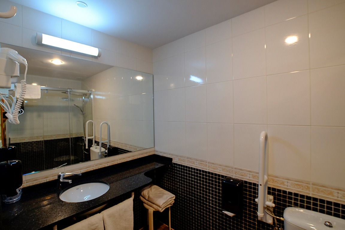 Baño con lavabo, espejo, inodoro, paredes de azulejos blancos y barra de apoyo.