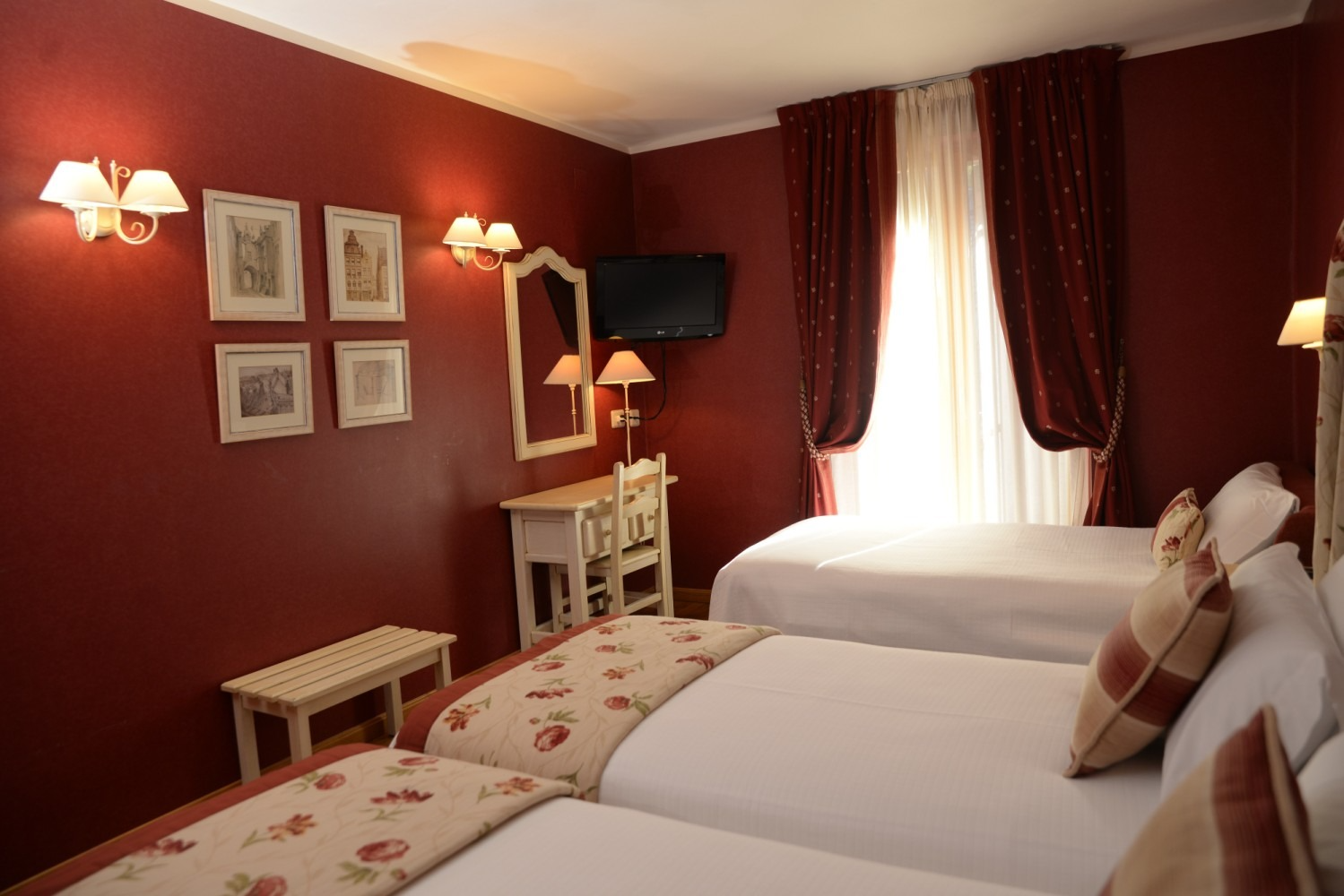 Habitación de hotel con tres camas, paredes rojas, molduras blancas, televisor pequeño, espejo y una ventana con cortinas .