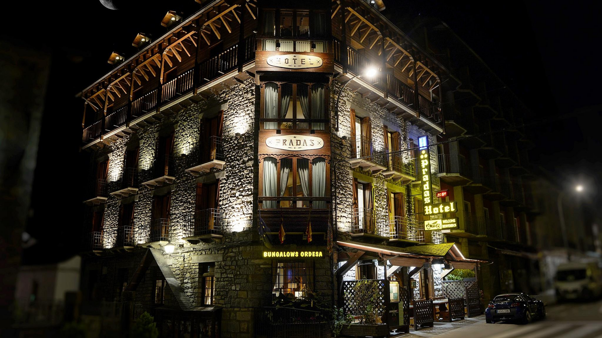 Edificio de piedra iluminado por la noche, cartel de hotel, coche aparcado fuera.
