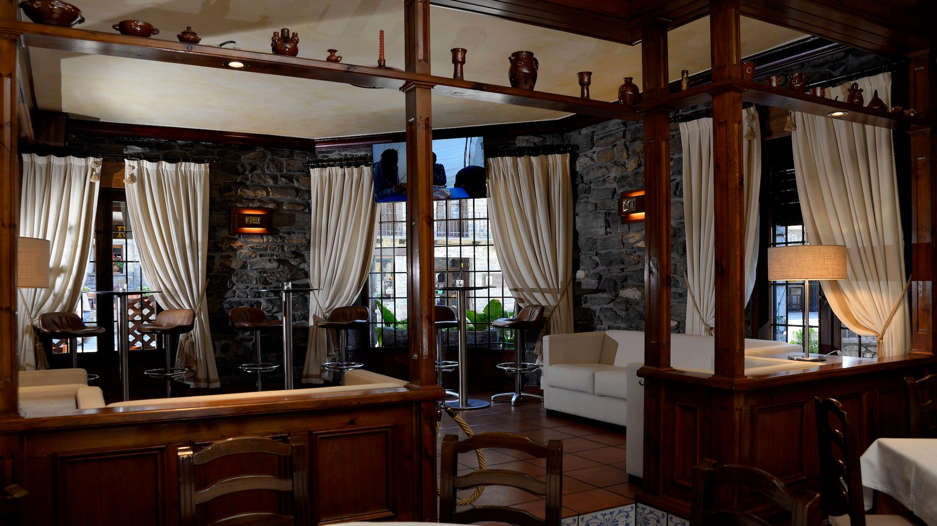 Interior de restaurante con molduras de madera oscura, paredes de piedra, cortinas y mesas de color crema.