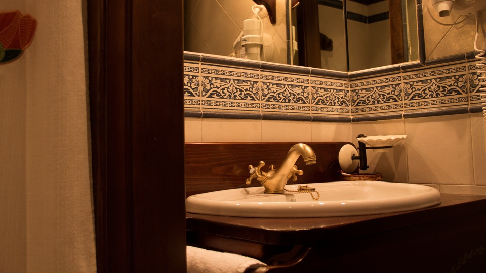 Lavabo de baño con grifería dorada, porcelana blanca, mueble de madera oscura y espejo enmarcado con azulejos estampados.