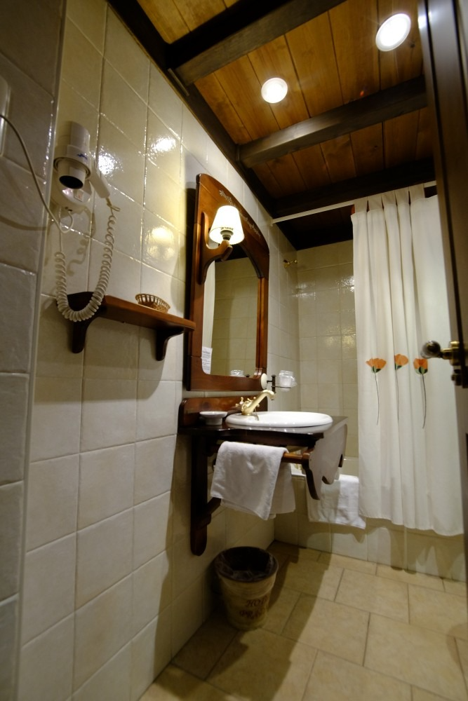 Baño con tocador de madera, espejo y paredes de azulejos. Hay toallas blancas colgadas y se ve una cortina de ducha.