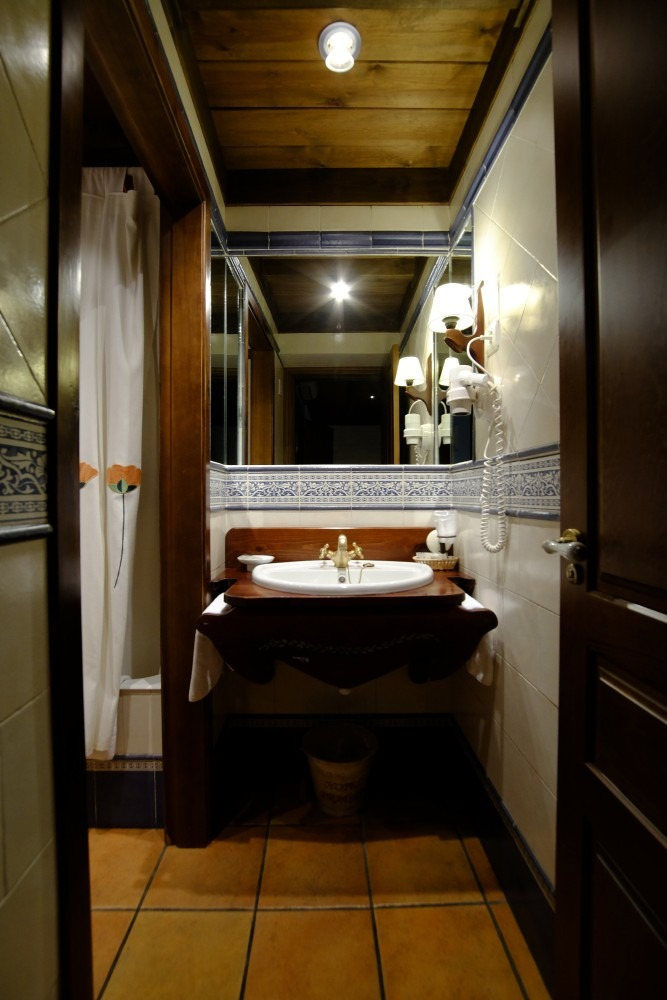 Baño con techo de madera y tocador. Un espejo refleja el interior, incluyendo una ducha tras una cortina.