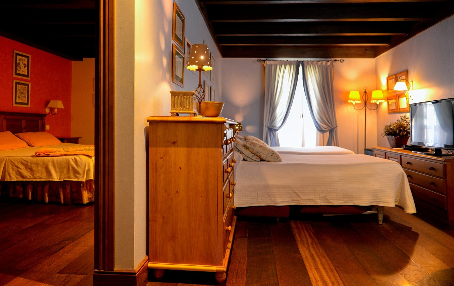 Dormitorio con suelo y muebles de madera, dos camas, TV y vista a otro dormitorio.