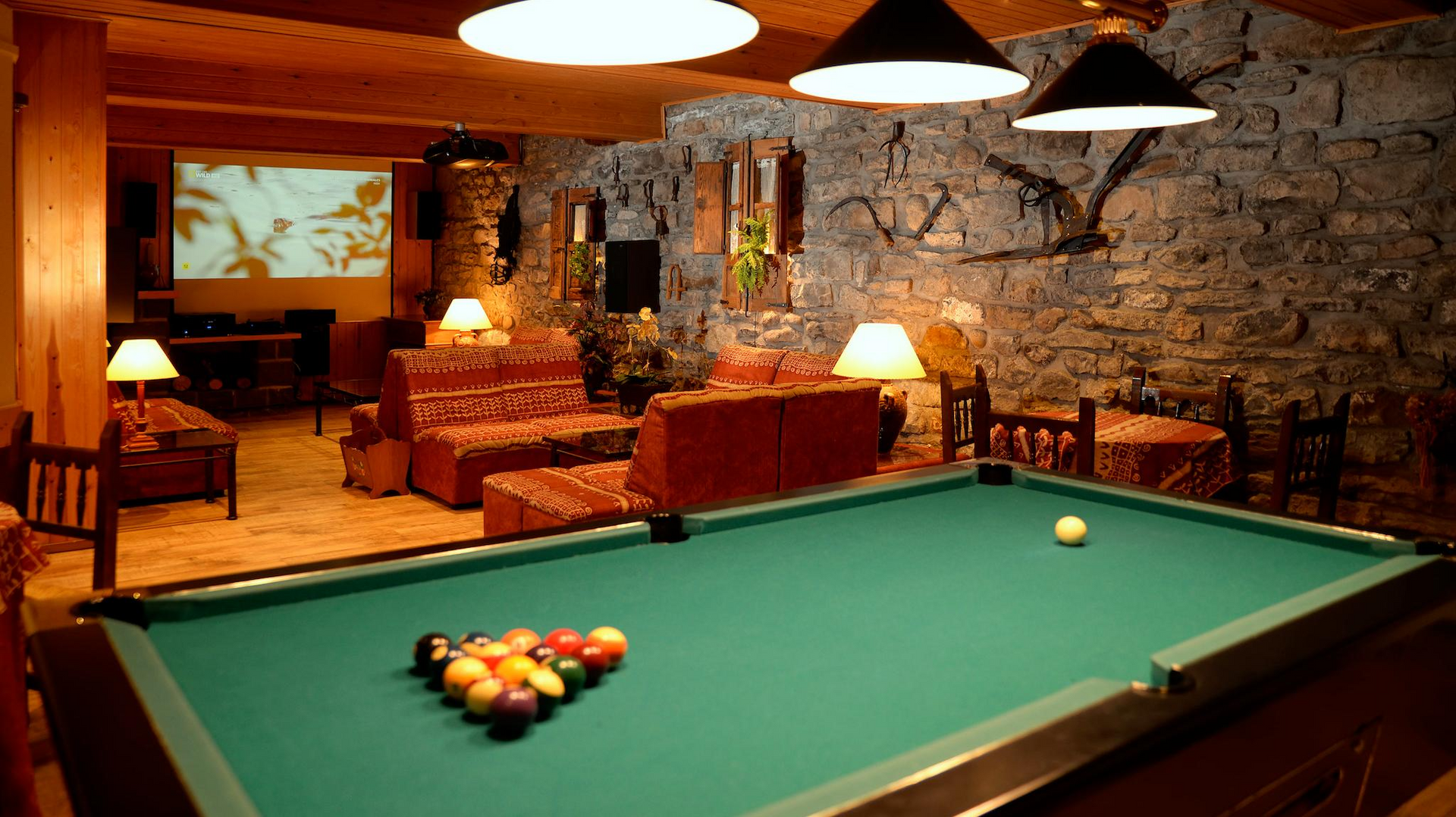 Mesa de billar en una habitación poco iluminada con paredes de piedra, sofás y pantalla de proyección.