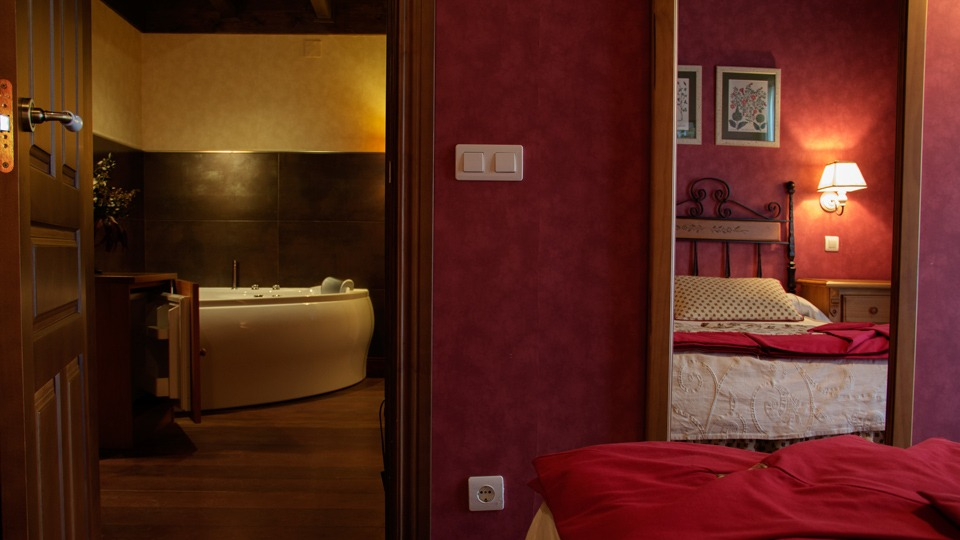 Dormitorio con una pared roja, un espejo que refleja la cama y una puerta a un baño con bañera.