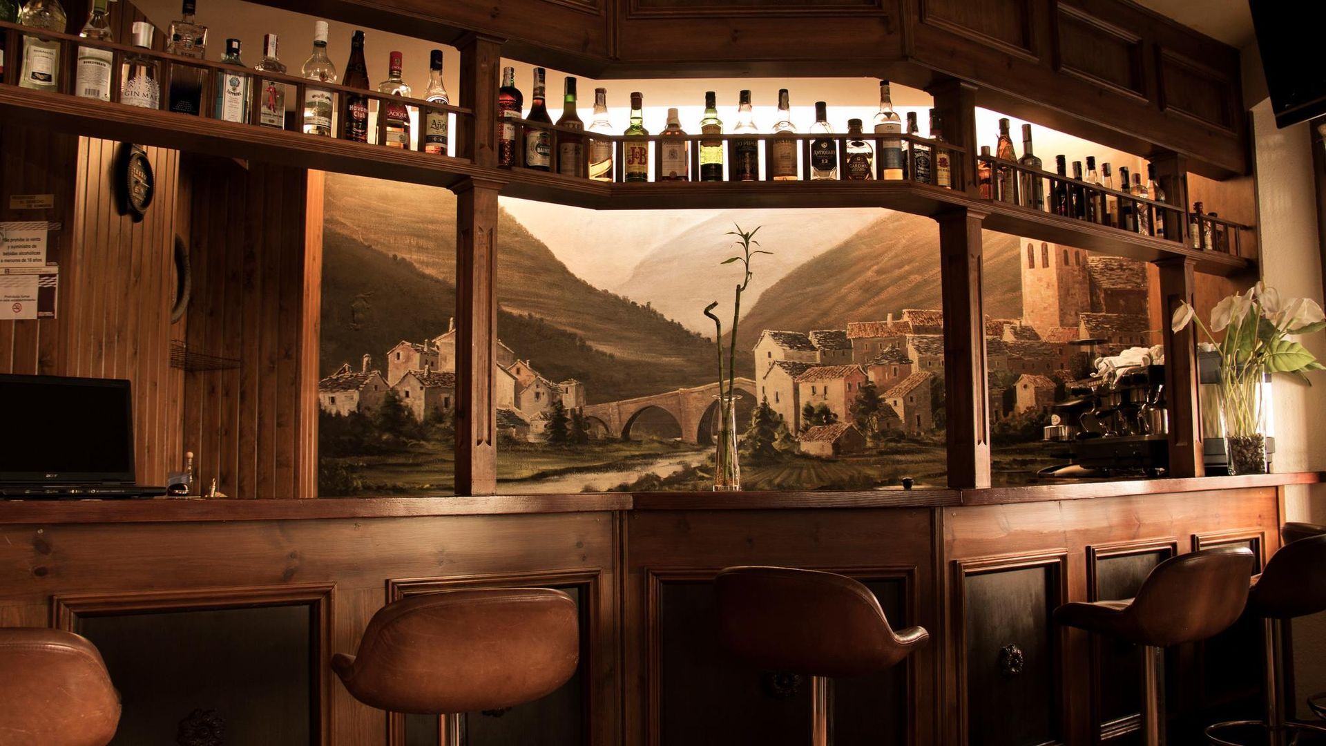 Interior de bar con paneles de madera, botellas en estantes, mural de paisaje y taburetes de bar.