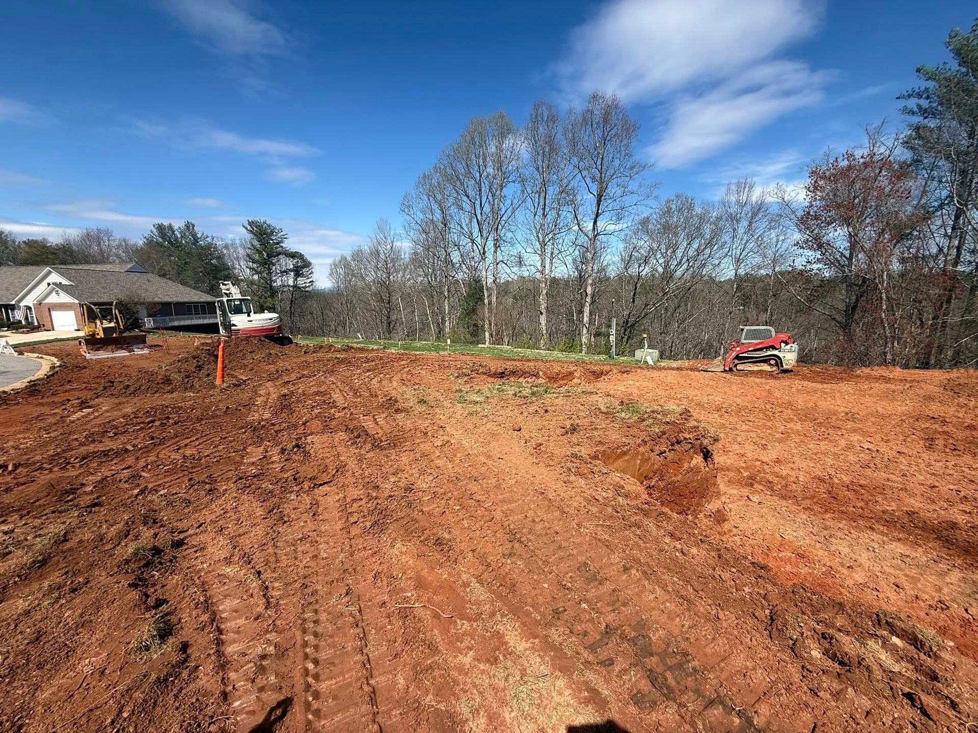 Wide dirt construction site with excavator, house at left, and trees under a blue sky