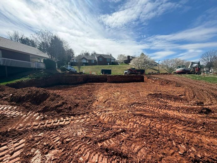 Cleared red dirt lot with tire tracks, houses and trees on the hillside under a blue sky