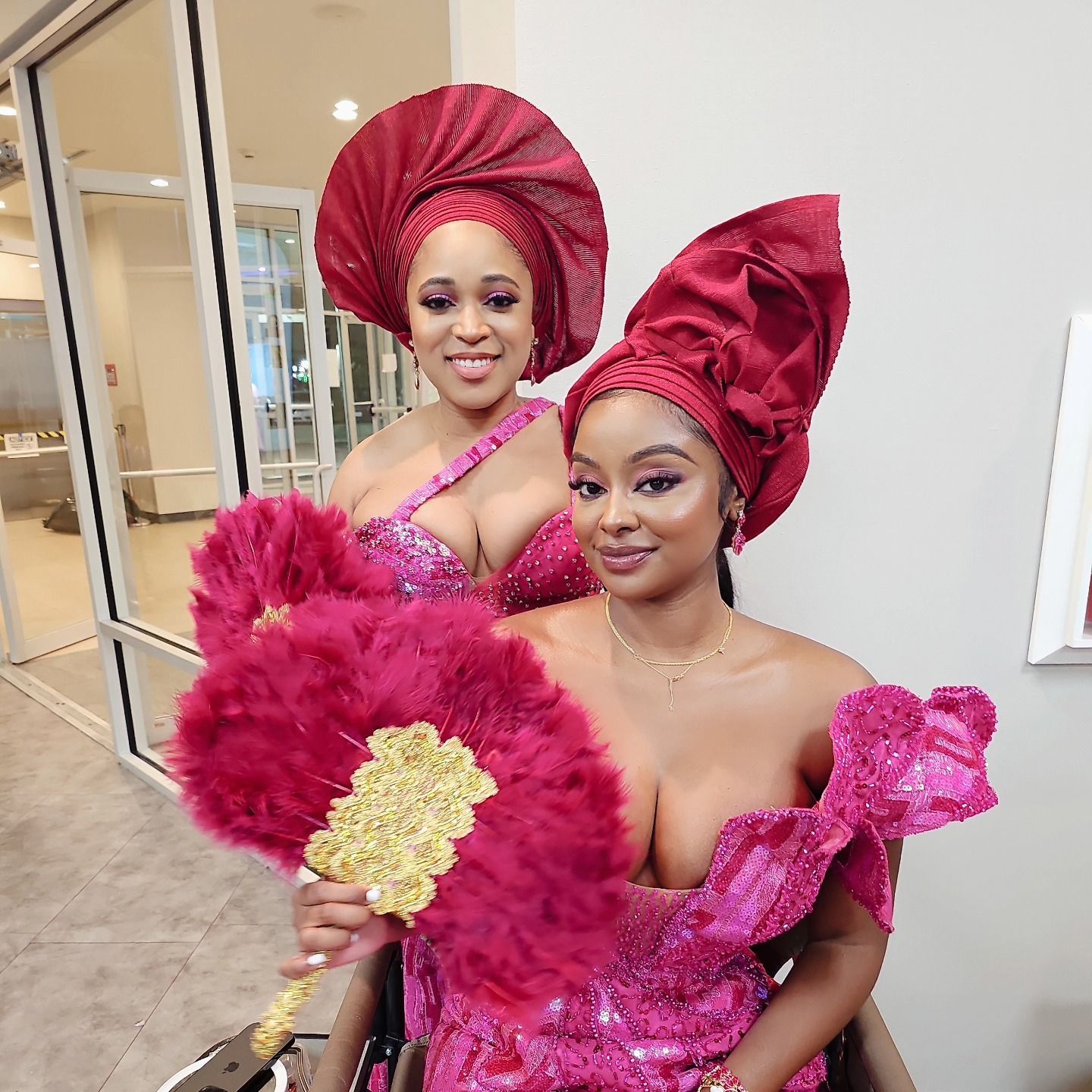 Two women in pink dresses and red turbans are holding pink fans.