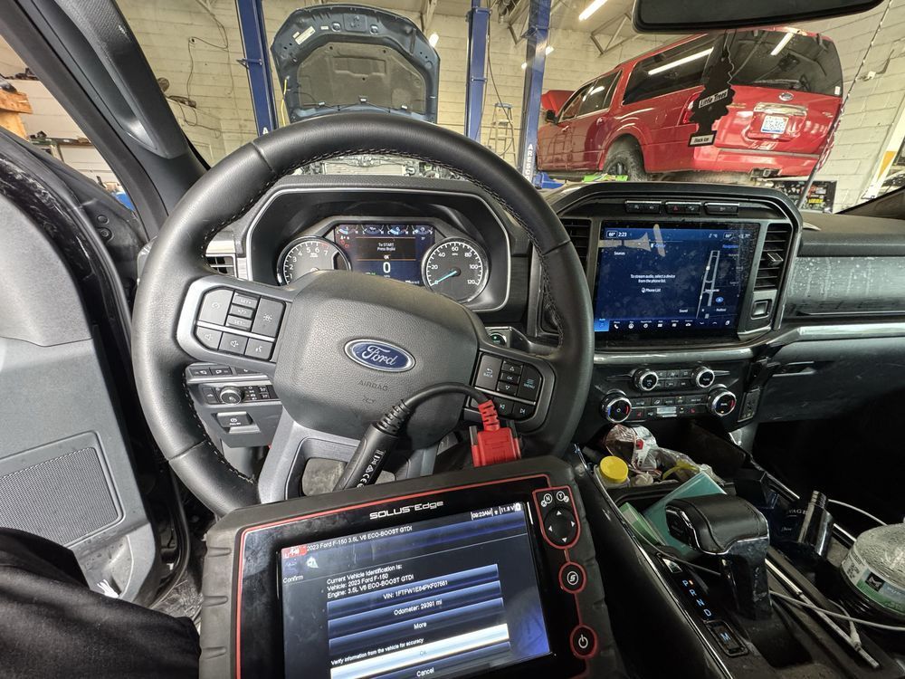 Car interior with steering wheel, diagnostic tool, and dashboard displays. A red truck is in the background.