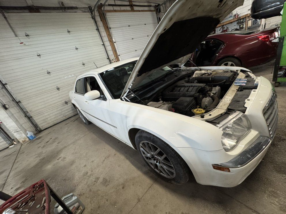 White Chrysler 300 with hood open in a garage, another red car in the background.
